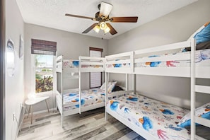 Een kamer met stapelbedden, een plafondventilator, een raam met jaloezieën en een klein tafeltje.