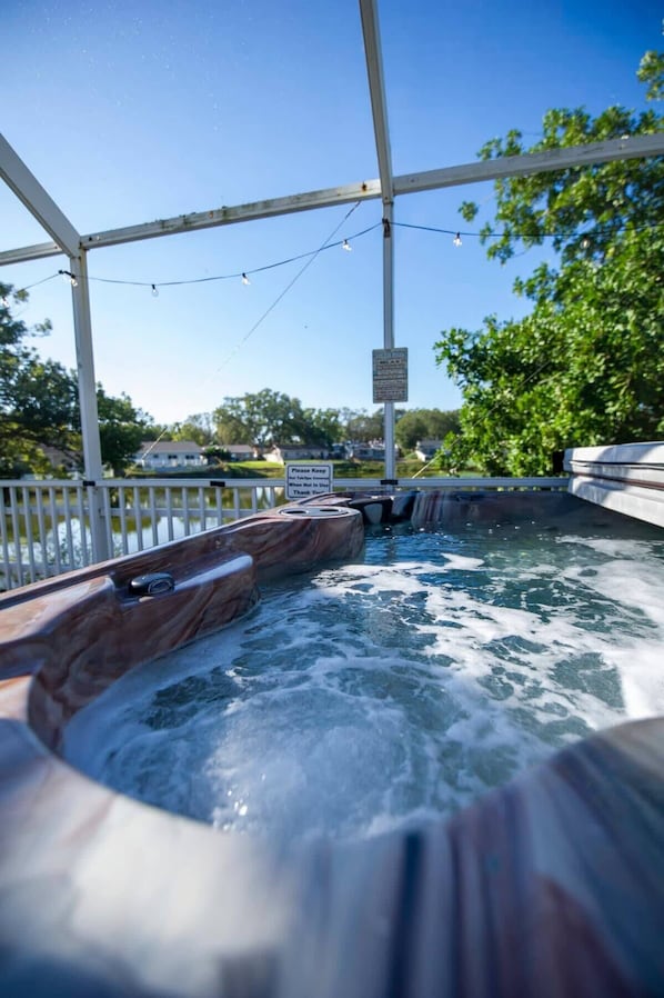 A hot tub area with a wooden deck and a metal railing, surrounded by trees and a clear sky. - Heated Pool + Spa Retreat-Minutes to Gulf Beaches (Palm Harbor)