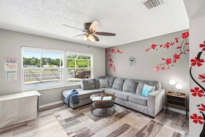Ein Wohnzimmer mit grauem Sofa, Couchtisch, Deckenventilator und Blick auf einen Pool durch ein großes Fenster.