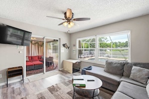 A spacious living room with a ceiling fan, a television, a sofa, a coffee table, and a large window offering a view of the outdoors.