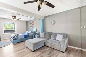 A modern bedroom with a bed, a grey sofa, a ceiling fan, and a window with a view. - Winter Escape • Heated Pool & Spa • Near Beach (Palm Harbor)