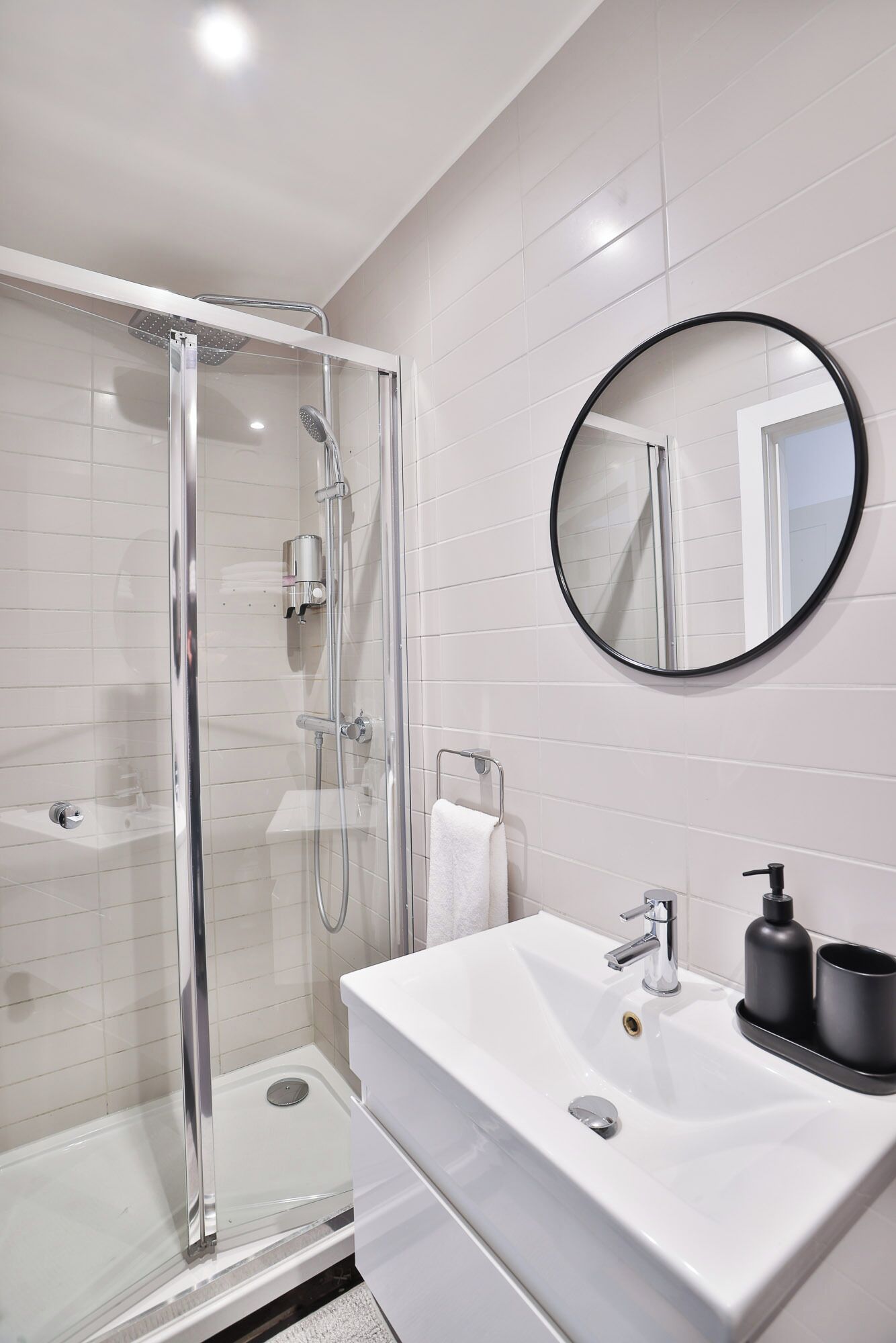 Un baño moderno con ducha de vidrio, un lavamanos blanco con dispensador de jabón negro y un espejo redondo en la pared.