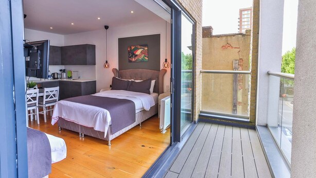 A modern bedroom with a large bed, a small kitchenette, and a balcony with a view of the city. - Goodwin (London)