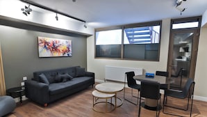 A modern living room with a grey sofa, a round coffee table, and a dining area with a table and chairs. A large abstract painting is on the wall, and there is a window with a view of a building outside. - Goodwin (London)