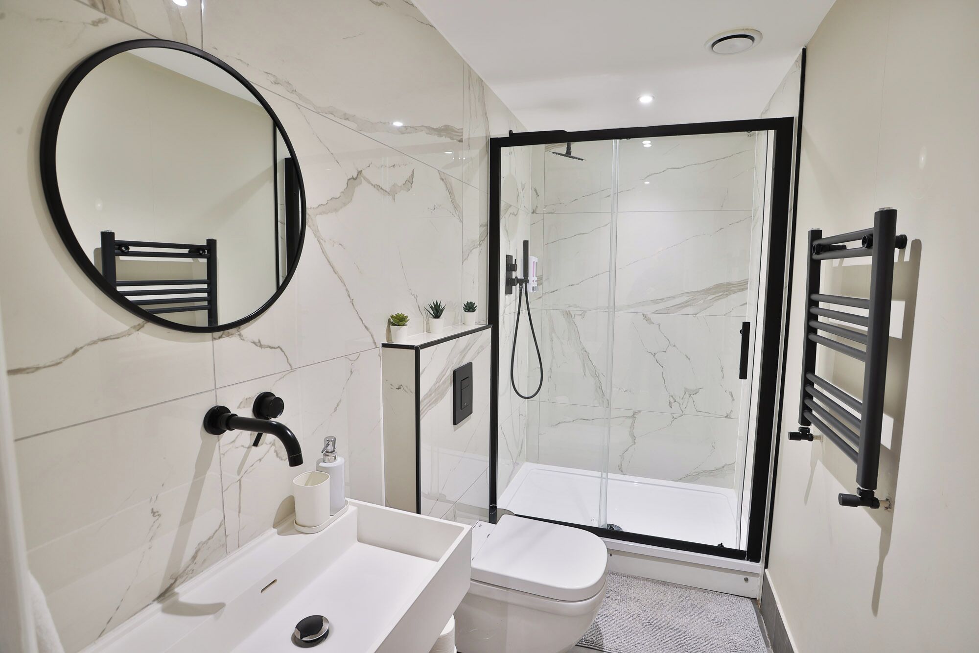 A modern bathroom with a white marble sink, a toilet, a glass-enclosed shower, and a black-framed mirror.