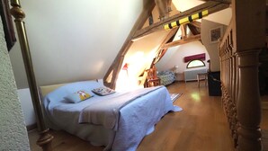 Een slaapkamer met een schuine kap, houten balken, een bed met een blauwe sprei, een nachtkastje en een raam met uitzicht.