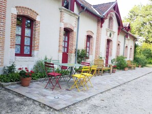 A building with a brick facade, red-framed windows, and a paved patio with outdoor seating. - Gîte Les Bambous, Sologne getaway, nature & relaxation 10 min from Chambord (Crouy-sur-Cosson)