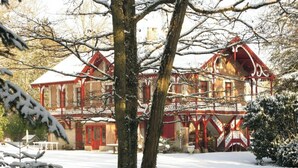 Een groot, rode baksteen gebouw met veel ramen en een balkon, omgeven door met sneeuw bedekte bomen en een heldere lucht.