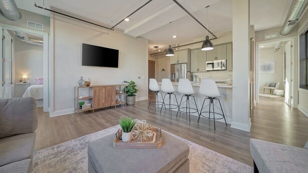 Een moderne woonkamer met een bank, een salontafel met decoratieve objecten, een aan de muur bevestigde televisie, en een keukenhoek met witte kasten en barstoelen.