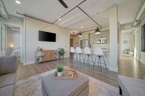 A modern living room with a sofa, a coffee table with decorative items, a TV mounted on the wall, and a kitchen area with white cabinets and bar stools.