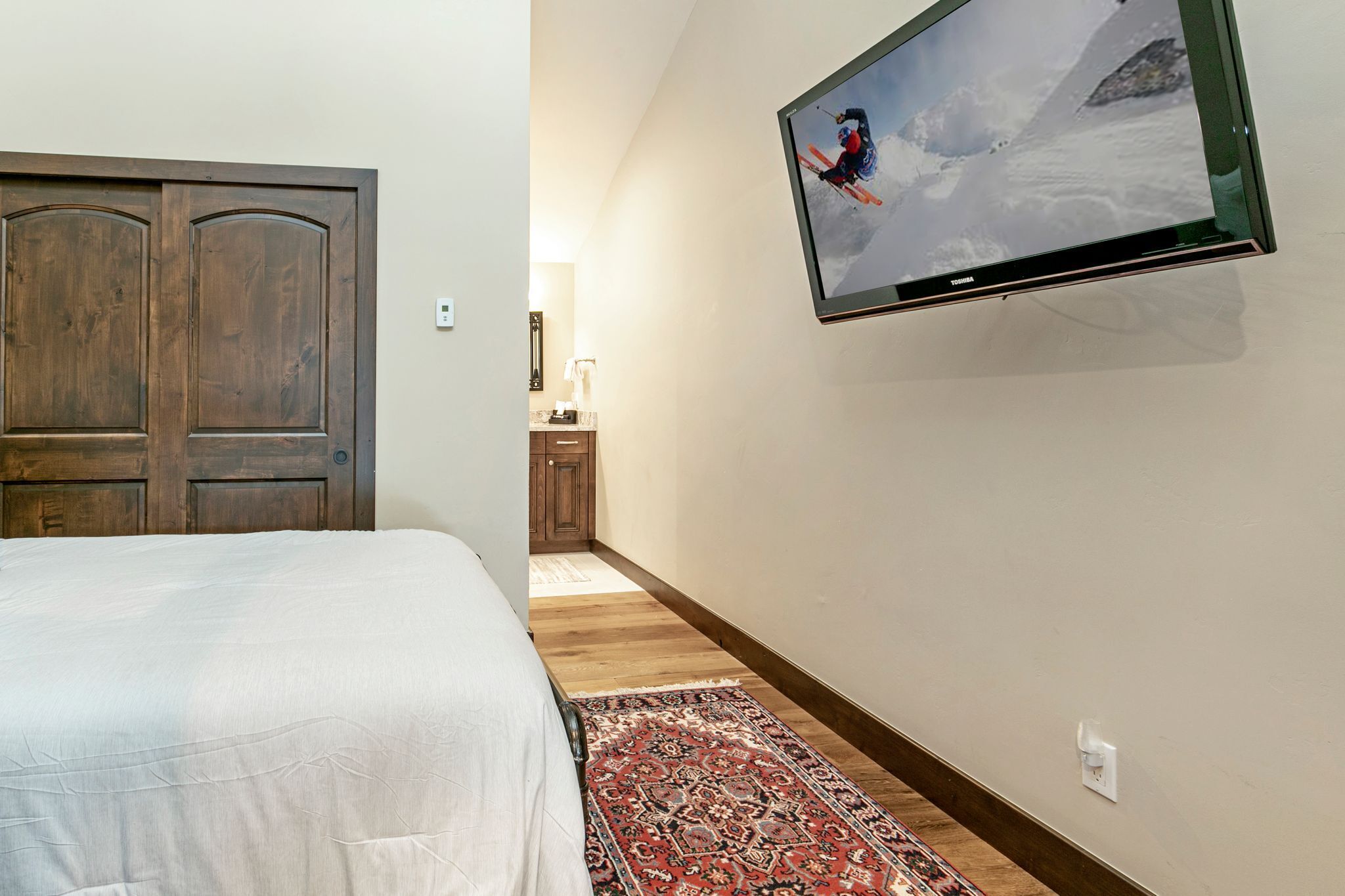 A bedroom with a bed, a TV mounted on the wall displaying a ski scene, a wooden door, and a hallway leading to a bathroom.