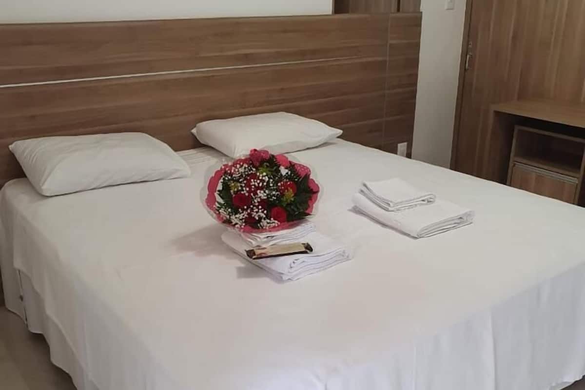 En snyggt bäddad säng med vita sängkläder, en bukett blommor och en hopvikt handduk på en träpanel som fungerar som sänggavel.