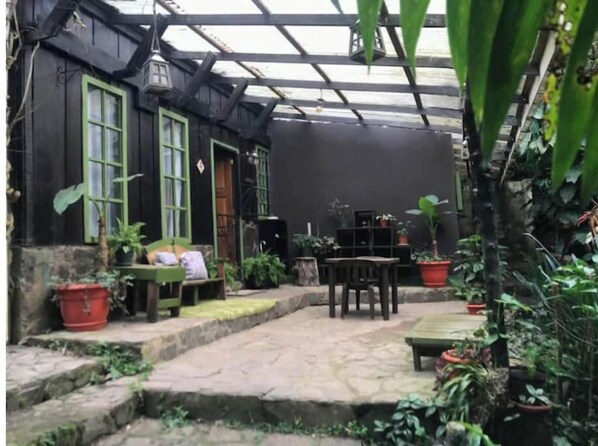 Relax in a serene garden setting with a wooden bench and outdoor dining table, surrounded by lush potted plants and a charming black-painted building.