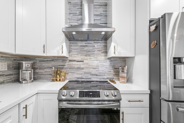 A modern kitchen with white cabinets, a stainless steel range hood, a built-in oven, and a refrigerator.