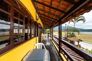 A covered outdoor area with a table and chairs, a view of a road and mountains, and a yellow building with large windows. - POUSADA ACQUA MARINA (Paraty)