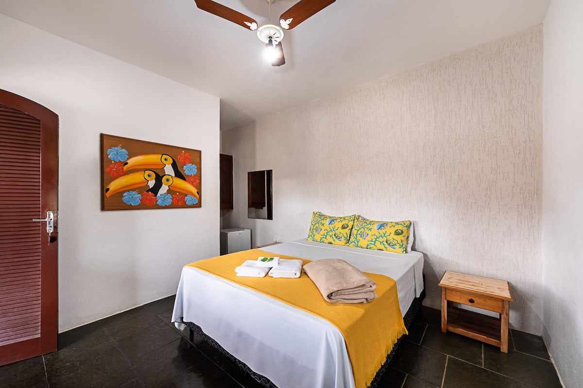 A hotel room with a bed, a wooden nightstand, a ceiling fan, and a painting of toucans.