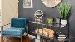 Ein modernes Wohnzimmer mit einem blauen Sessel, einem Glastisch mit Dekoration, einem runden Spiegel und einer Zimmerpflanze.