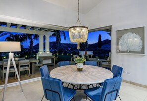 A dining area with a round table, blue chairs, a chandelier, and a view of mountains and palm trees.