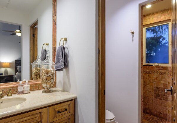 A bathroom with a large mirror, a sink with toiletries, a glass container with bath products, and a shower area with a window.