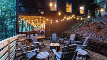 An outdoor seating area with Adirondack chairs and a round table, surrounded by a wooden deck and string lights.