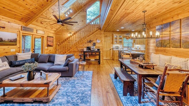 A cozy log cabin interior with a wooden ceiling, a sofa, a coffee table, a dining table, and a kitchen area. - Ellijay River Access (Trout!) | Hot Tub| Firepit | Games | Dogs Welcome! (Cherry Log)