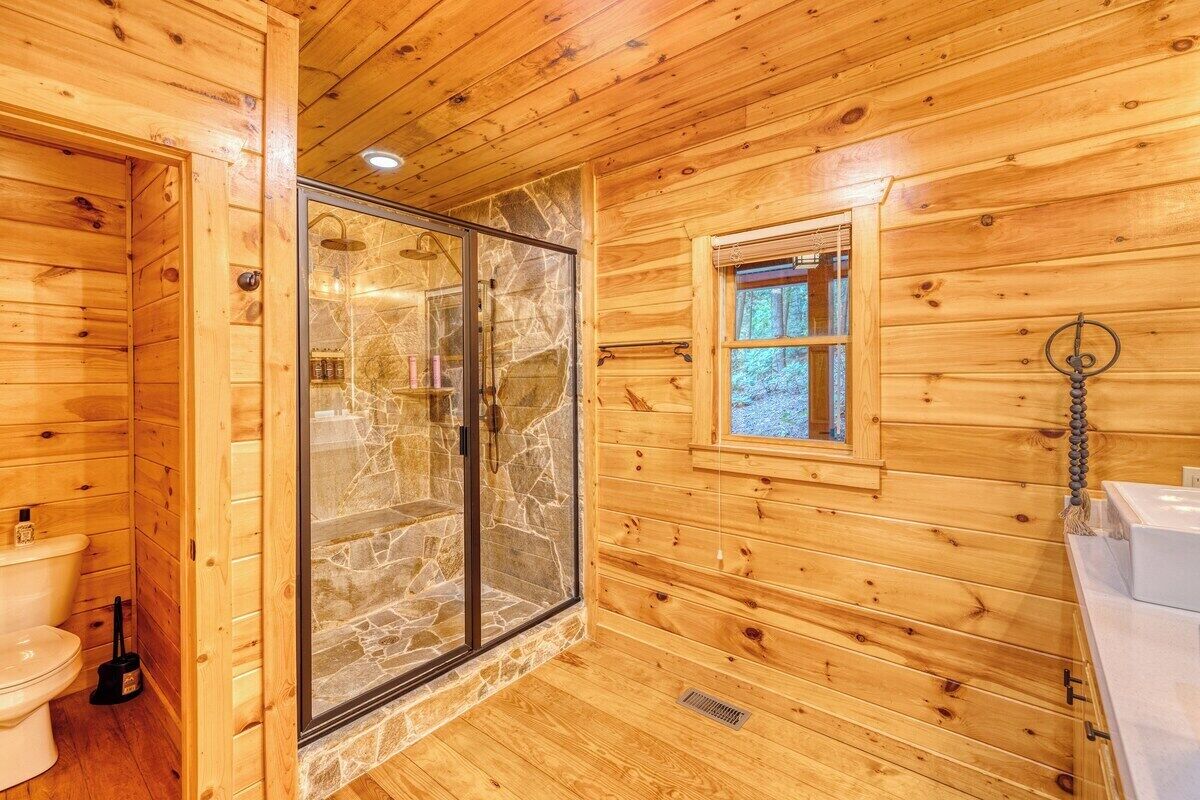 A wooden cabin bathroom with a stone shower enclosure, a toilet, and a window with a view of the outdoors.