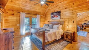 A wooden cabin bedroom with a bed, a nightstand, a ceiling fan, and a painting of a river scene. - Ellijay River Access (Trout!) | Hot Tub| Firepit | Games | Dogs Welcome! (Cherry Log)