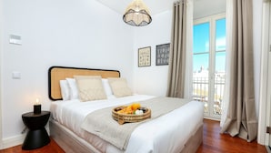 Una cama bien hecha, con una bandeja de frutas y una vela, un lámpara colgante, y vista a un balcón a través de una ventana.