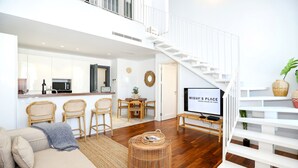 Uma sala de estar ampla com um sofá, uma mesa de centro e uma televisão em primeiro plano. Uma escada com corrimão branco leva a um andar superior.