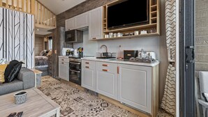 A modern kitchen with white cabinets, a large TV, and a dining area with a patterned rug. - The Escape - Luxury Pod, Close to Beach (Wisemans Bridge)