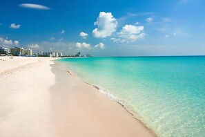 A beach with clear turquoise water, a sandy shore, and a city skyline in the background.