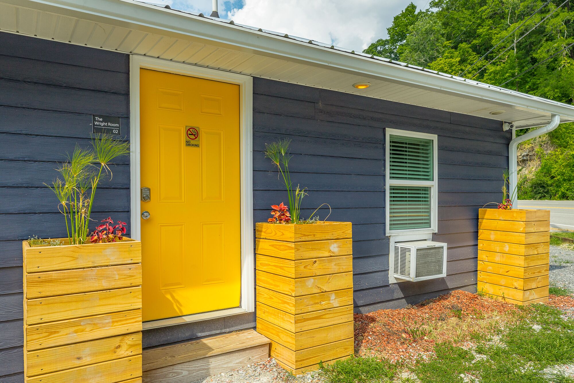 A house with a yellow door, a window with green shutters, and wooden planters with plants.