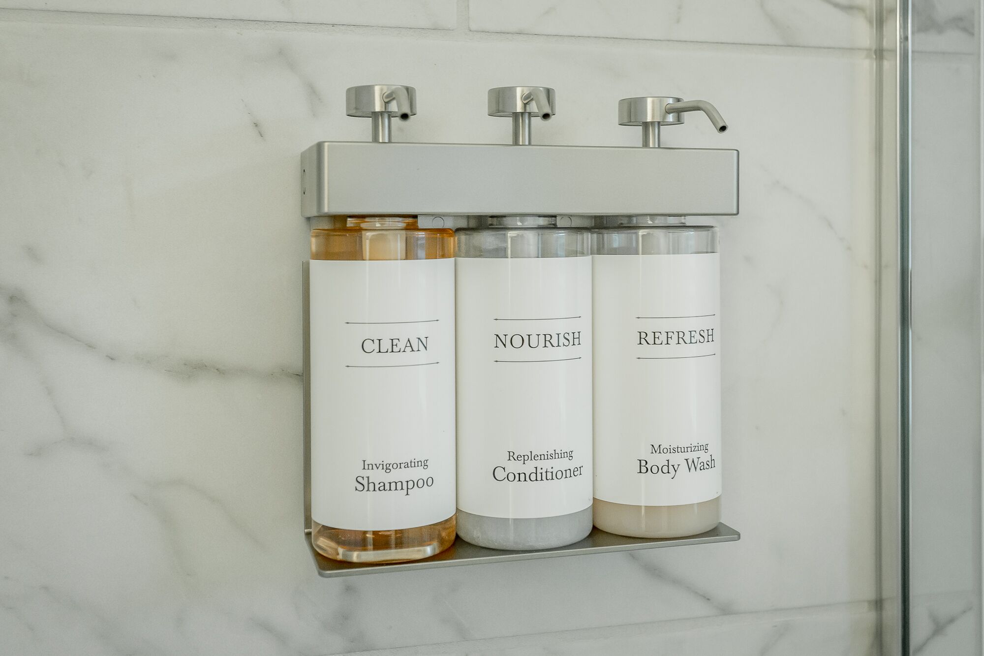 A bathroom with a marble wall and a shelf holding three bottles of shampoo, conditioner, and body wash.