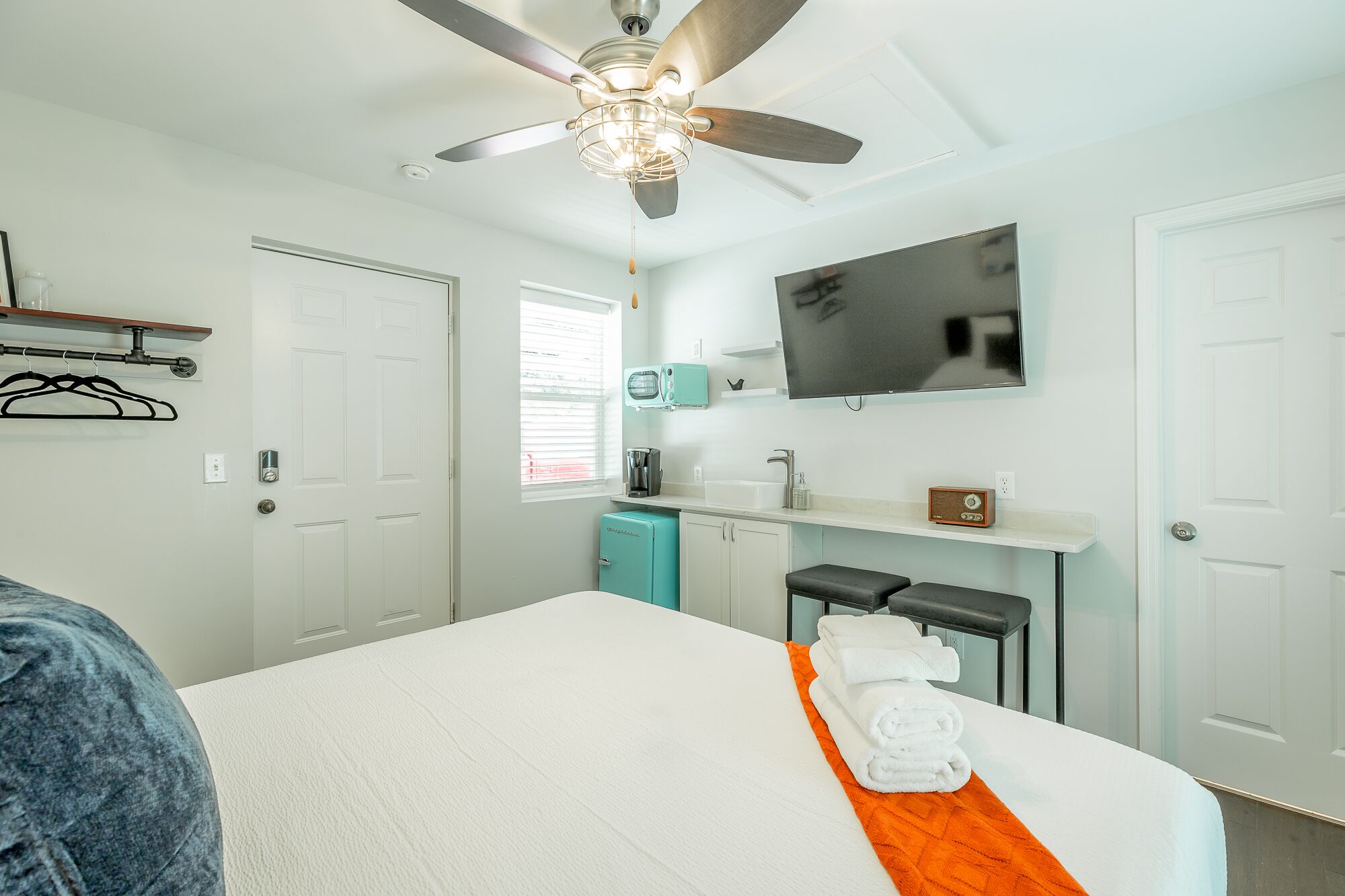 Une chambre d’hôtel avec un lit, un ventilateur de plafond, une télévision, une petite kitchenette et une fenêtre.