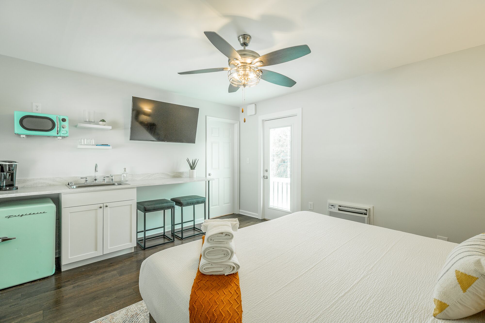 A modern bedroom with a bed, a ceiling fan, a TV, and a kitchenette.
