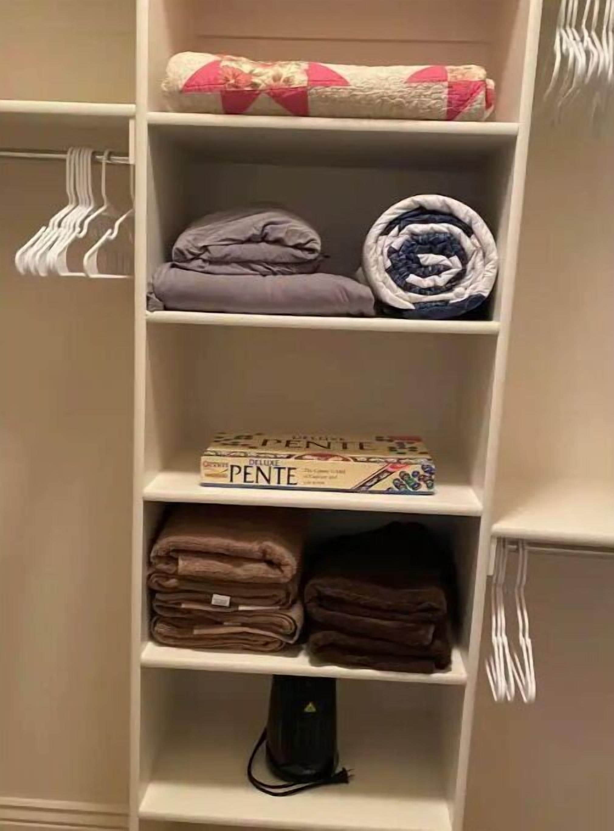 Ein Kleiderschrank mit Regalen, der gefaltete Kleidung, eine mit 'PENTE' beschriftete Kiste und eine Mehrfachsteckdose beinhaltet.