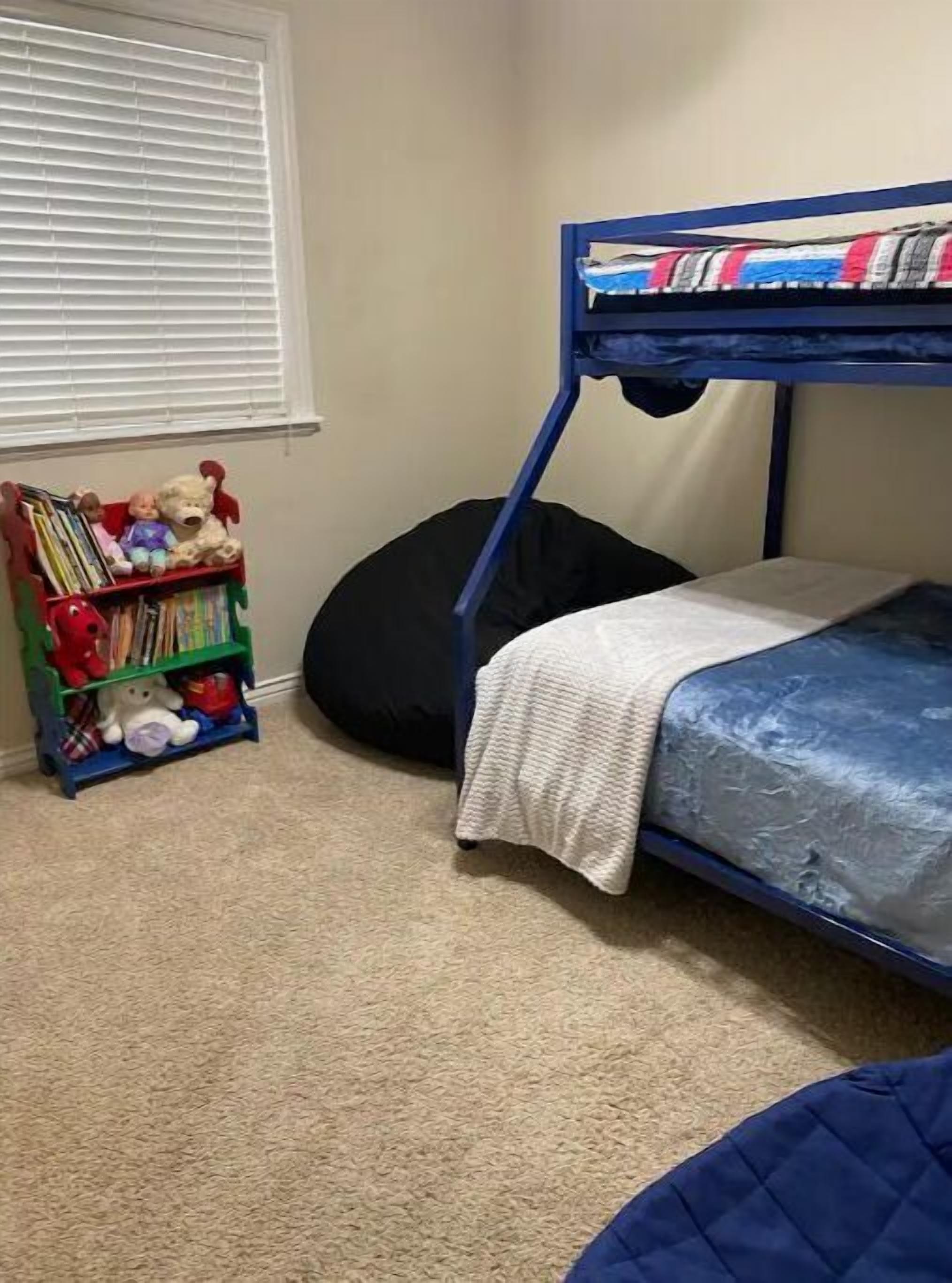 A bunk bed with a white blanket, a bookshelf with toys and books, and a black bean bag chair in a room with beige carpet.