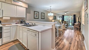 A kitchen with white cabinetry, a dishwasher, and a sink. There is a dining area with a table and chairs. - True Blue Golf Resort Condo - 3 Mi to Beach! (Pawleys Island)