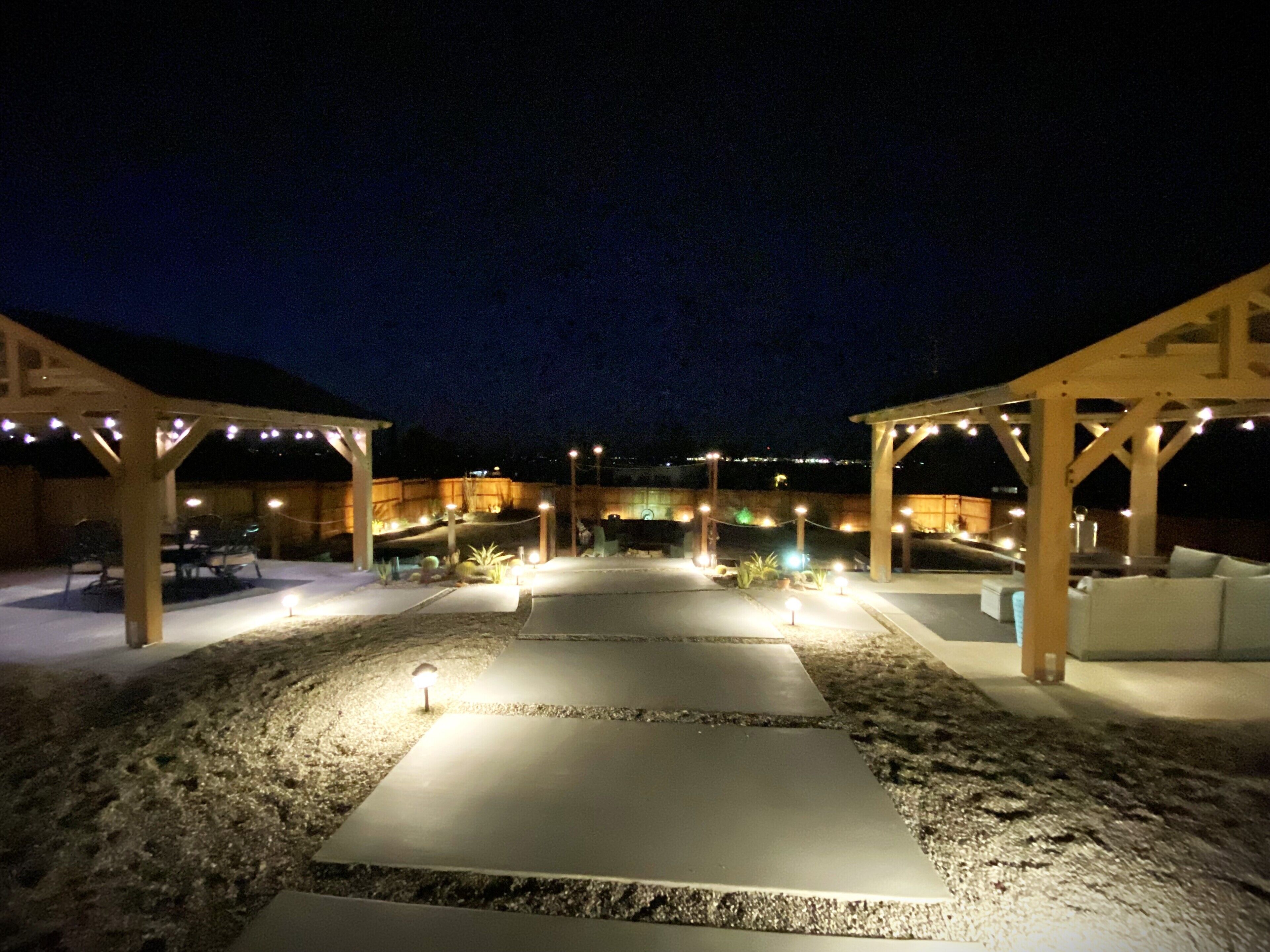 An outdoor patio area with a concrete pathway, wooden pergolas, string lights, and outdoor furniture.