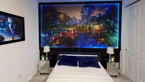 Camera da letto con una grande TV a parete che mostra una vivace scena di paesaggio. Sono presenti due comodini con lampade, un letto con cuscino blu e un quadro appeso alla parete.