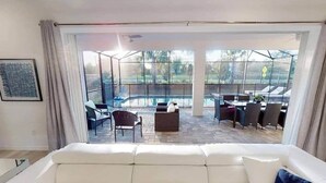 Un soggiorno moderno con un divano bianco, un'area pranzo con sedie e un tavolo, e vista su una piscina attraverso ampie vetrate.