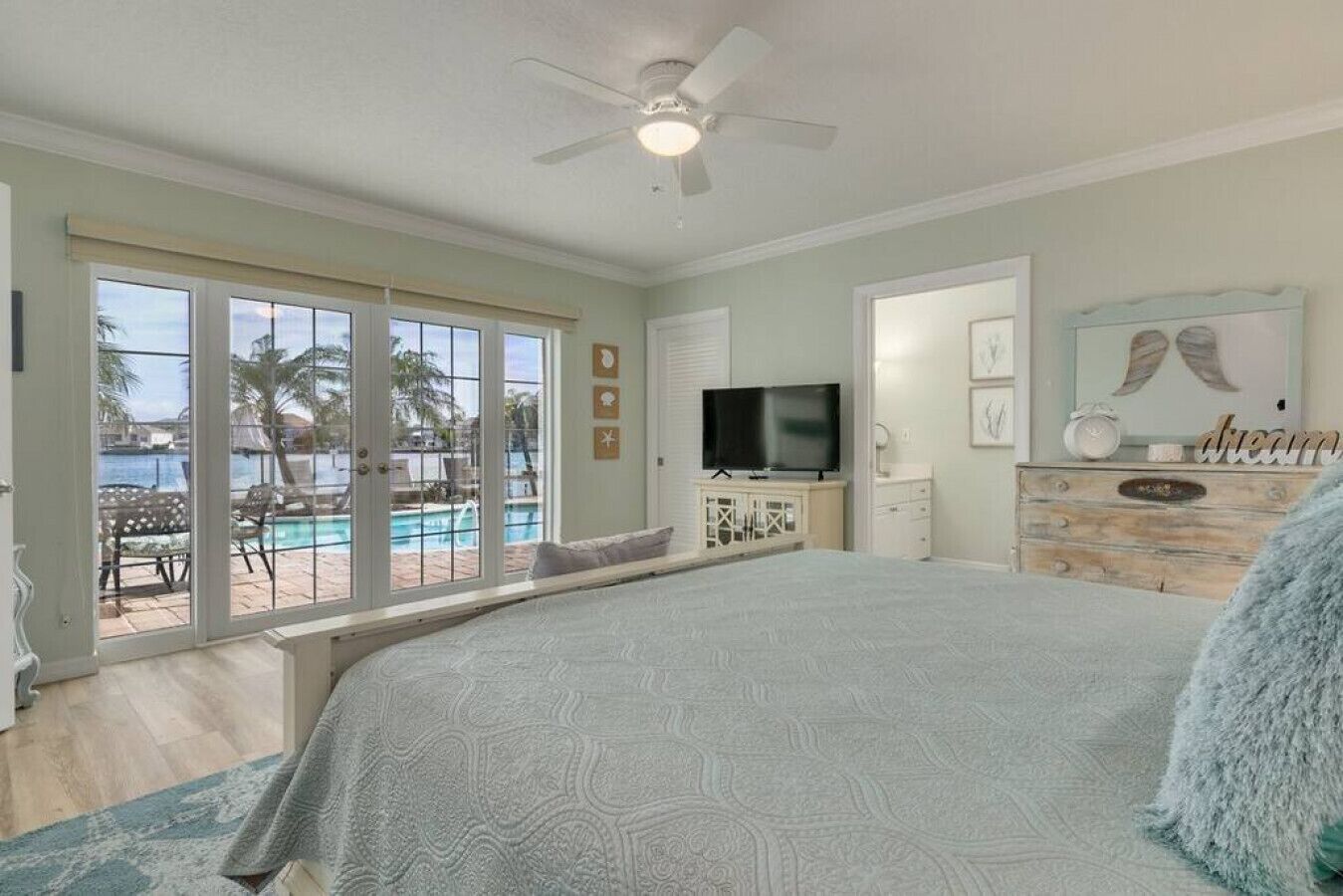 A bedroom with a large bed, a TV, a dresser, and a view of a pool and palm trees.