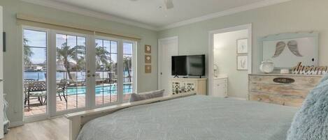 A bedroom with a large bed, a TV, a dresser, and a view of a pool and palm trees.