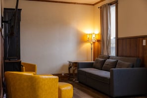 Ein Wohnzimmer mit einer Couch, zwei gelben Sesseln, einem kleinen Tisch, einer Lampe und einem Fernseher.
