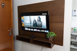 Litteänäyttöinen televisio on kiinnitetty puiseen seinään, ja sen mukana on kaukosäädin. Sen alapuolella hyllyllä on pieni ruukkukasvi.