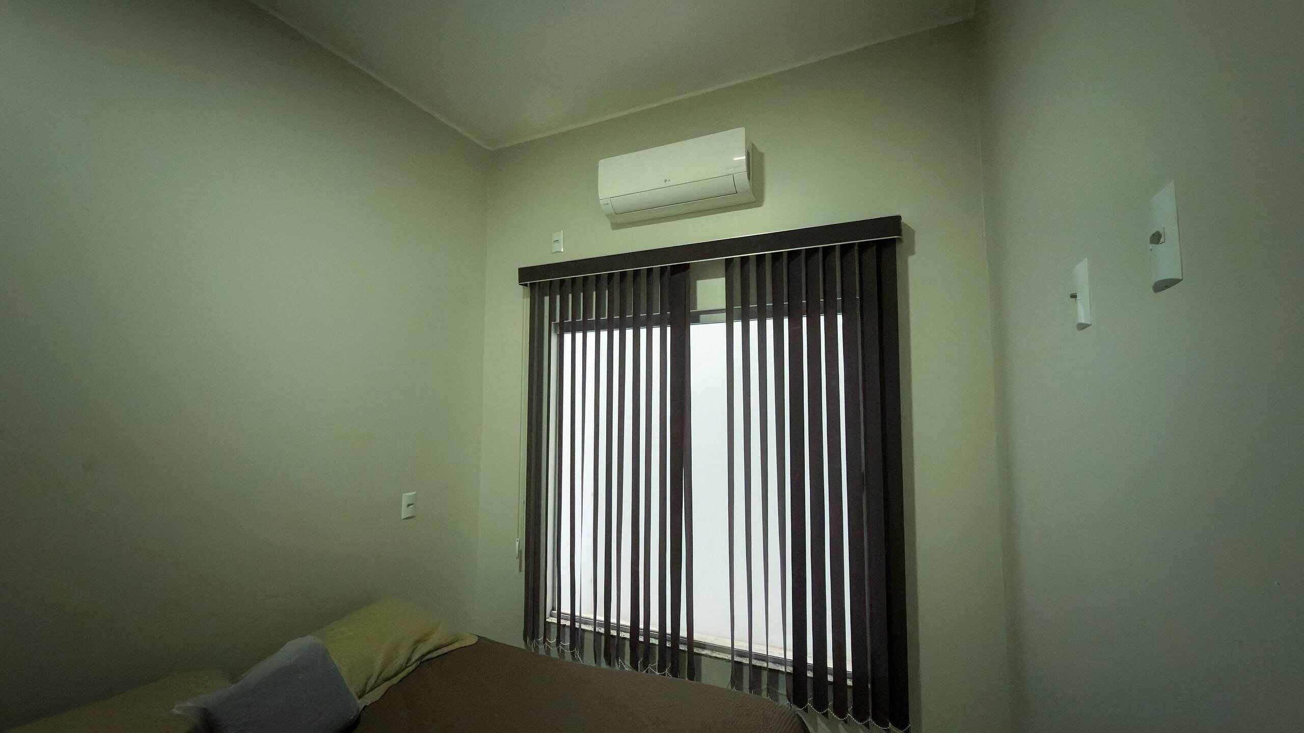 A room with a bed, a window with vertical blinds, and an air conditioning unit on the wall.