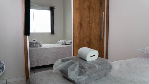 A room with a bed, a roll of toilet paper on the bed, a fan, and a window with a curtain. - CONDOMÍNIO SONHARE - 104 (Anapolis)