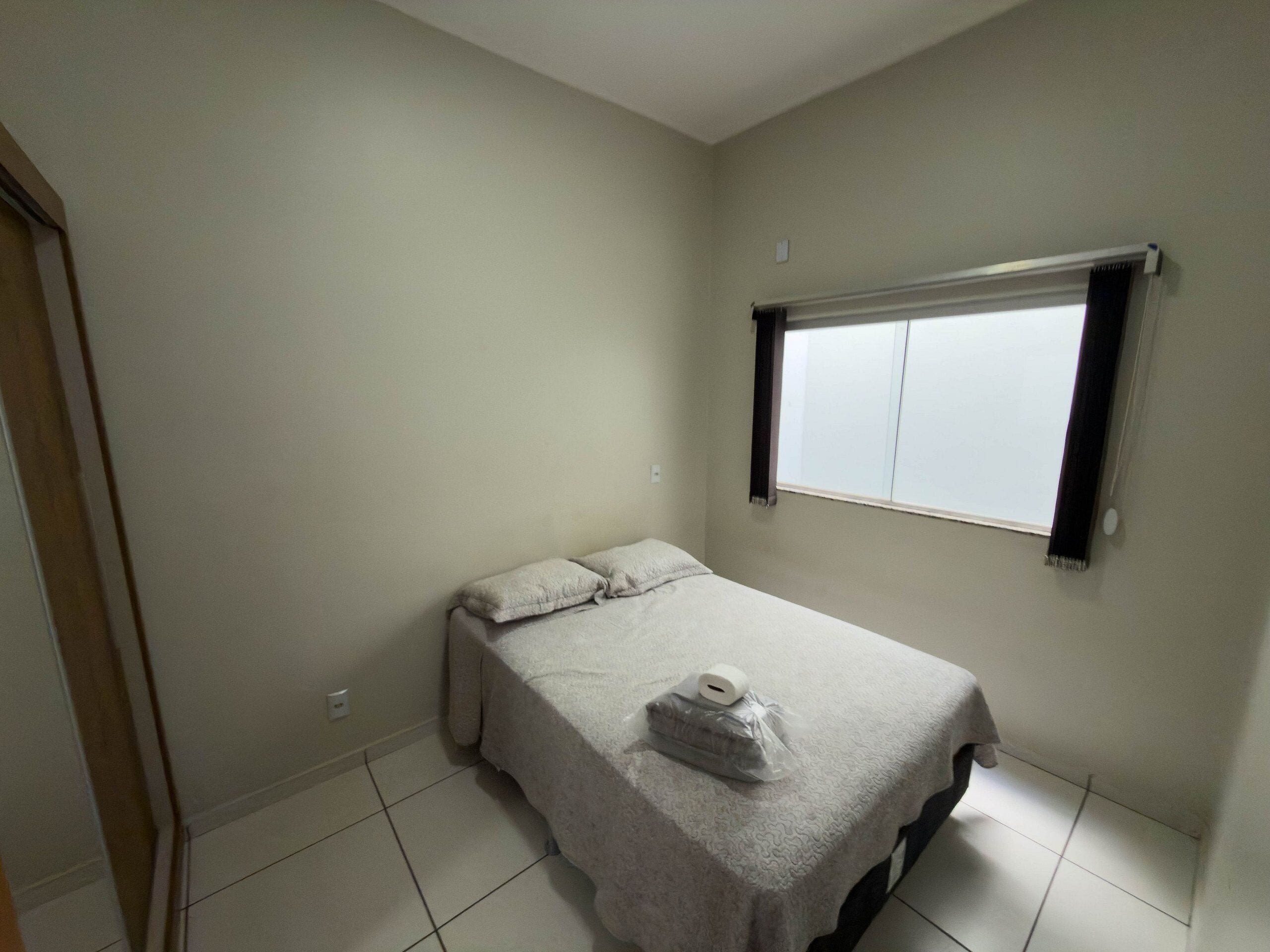 Ein kleines Zimmer mit einem Einzelbett, einem Fenster, einer Decke auf dem Bett und einer Toilettenpapierrolle.