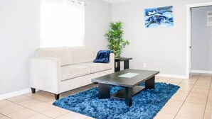 A living room with a white sofa, a coffee table, a blue rug, a plant, and a painting on the wall. - Modern Style Minimalistic Home! (Dallas)