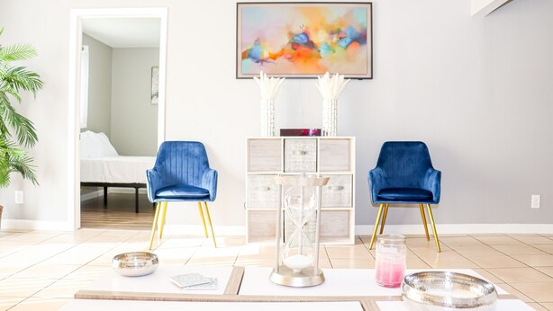 A modern living room with a bed in the background, two blue chairs, a glass hourglass, and a painting on the wall. - Modern Style Minimalistic Home! (Dallas)